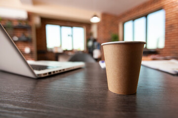 Closeup of disposable mug placed on vacant desk with laptop nearby, highlighting clean brick wall workspace. Coffee cup on table near digital device, set in startup office with no people.