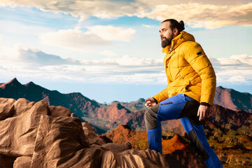 Naklejka premium Male trekker takes in the breathtaking mountain landscape on rocks, enjoying the scenic background, crisp air and rugged peaks. Excitement of discovering new horizons for wellbeing.