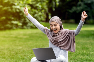 Emotional muslim girl looking at laptop screen, screaming happily and raising hands up, got great news, resting at park, empty space