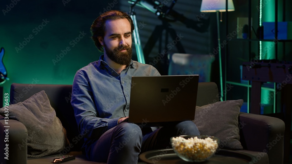 Poster man typing on laptop keyboard in neon lit apartment, sitting on couch and watching tv with bowl of p