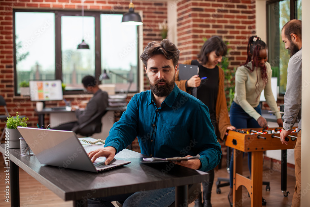 Poster bearded businessman focused on reading documents and entering findings on laptop in modern office. m