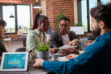 Diverse coworkers collaborate on project in modern workspace with documents and devices. Team...
