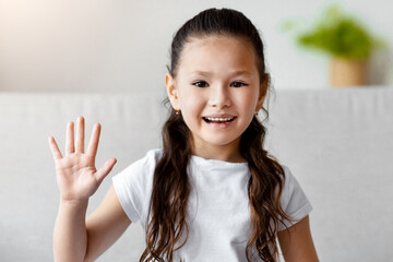 Hello. Cute Asian Kid Girl Waving Hand Smiling To Camera Sitting On Sofa At Home
