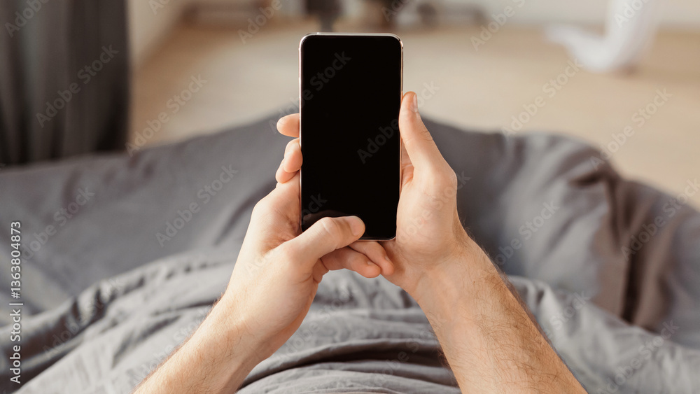 Poster unrecognizable guy using mobile phone with blank screen lying in bed at home. panorama, mockup, pov
