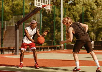 Two millennial sportsmen playing basketball at outdoor arena on warm summer day