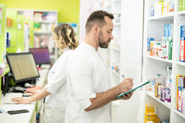 Obraz premium A focused pharmacist records product details while a colleague organizes items on shelves, reflecting teamwork in pharmacy management.