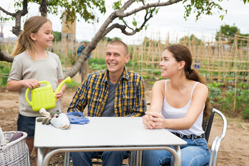 Portrait of cheerful young man, woman and little girl sitting at table resting after gardening at...