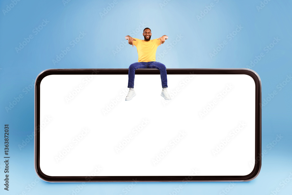 Poster smiling young african american man sitting on top of giant cellphone, pointing down at empty screen 