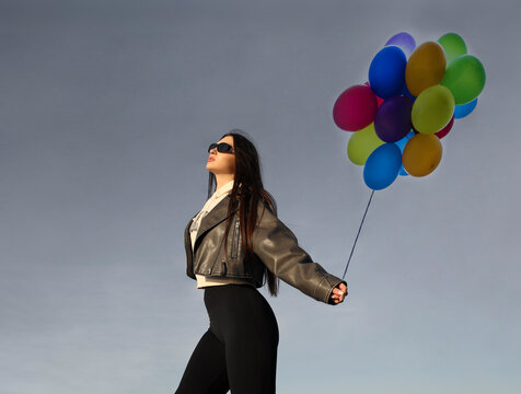young latin latino hispanic brunette woman in short stylish black leather jacket and white hoodie hold bunch of balloons. relaxed female lady girl against the blue clear sky. hands up fly abone land
