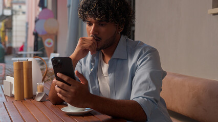Indian man Arabian male guy student tourist traveler customer in cafe city drink coffee thoughtful businessman choose travel tickets service app internet order pay online shopping mobile phone outdoor