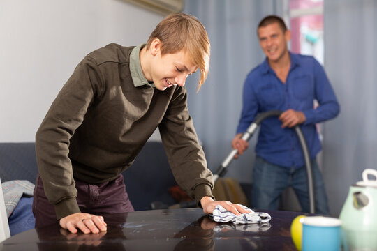 Positive teenage boy cleaning table with rag, man vacuuming the sofa at home