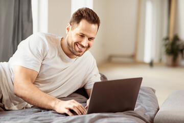Happy Freelancer Man Using Laptop Computer Browsing Internet Working Online Lying In Bed In Bedroom At Home. Distant Job, Web Freelance Career Lifestyle Concept