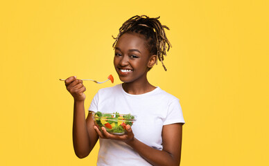 Smiling black girl eating fresh salad over yellow studio background, healthy food and diet concept