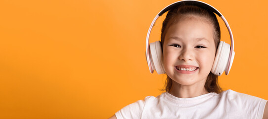 Closeup headshot of cute little asian girl listening to music wearing wireless headset with smile,...
