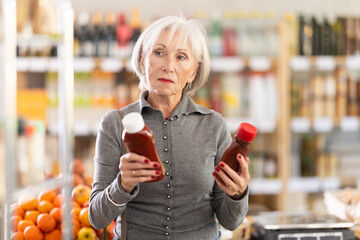 Fototapeta premium Elderly woman buyer choosing tomato ketchup sauce in grocery store