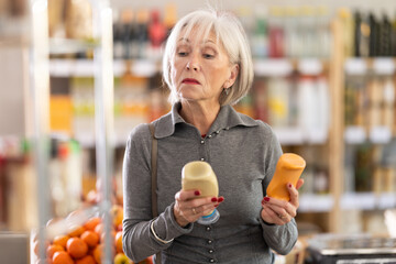 Elderly woman buyer choosing mustard or mayonnaise sauce in bottle in grocery store