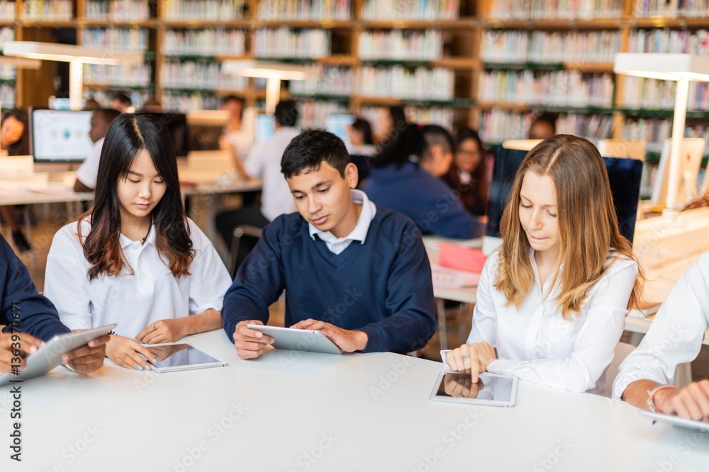 Fototapete Students studying in a library, using tablets. Diverse group focused on learning. Library setting with books, students, and technology. Educational environment. Diverse students learning at library.