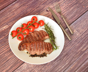 Two pieces of grilled beef fillet with cherry tomatoes, rosemary and cutlery on a dark wooden table.