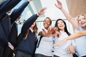 Fototapeta premium Group of diverse students in uniforms, smiling and raising hands. Diverse students, diverse smiles, diverse energy. Happy students, happy moments. Happy students stacking hands in teamwork and unity.