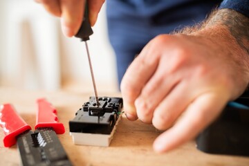Close-up of hands using a screwdriver on an electrical component, with wire strippers nearby on a wooden surface. Focus on electrical work and tools. Hand working on electrical material.