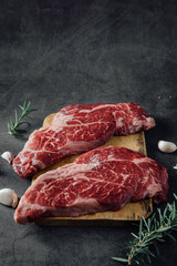 Fresh Raw Beef Steaks on Wooden Board with Garlic and Herbs on Dark Background