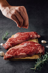 Hand Sprinkling Salt on Fresh Raw Meat Cuts with Herbs on Dark Background