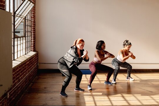 Three women exercising indoors, performing squats. Fitness workout with diverse women in athletic wear. Group exercise in a sunlit room, focusing on health. Healthy women exercising at fitness gym.