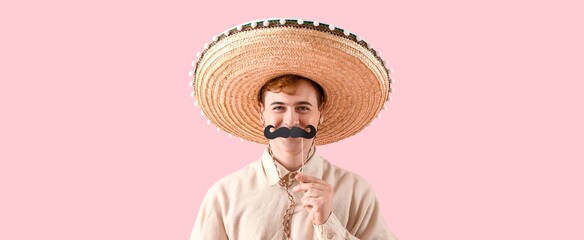 Funny young man in sombrero and with paper mustache on pink background