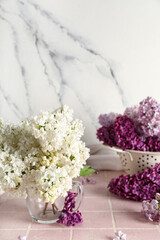 Obraz premium Colander and cup of beautiful fragrant lilac flowers on pink tile near grunge white background