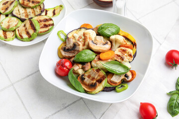 Plates with grilled vegetables and basil on white tile background