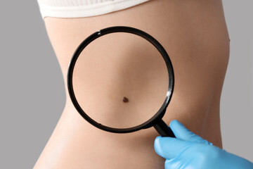 Dermatologist with magnifier examining mole on woman's belly against light background, closeup