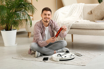 Young man with mobile phone changing brush of modern robot vacuum cleaner at home