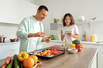 Young couple with recipe peppering cut bell peppers on baking tray in kitchen