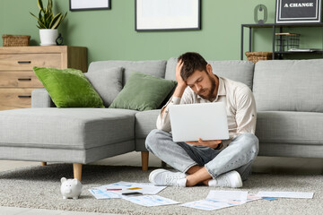 Young man in debt using laptop on carpet at home