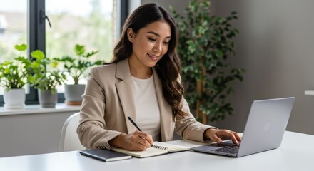 Professional woman working at desk with laptop and notebook in modern office setting