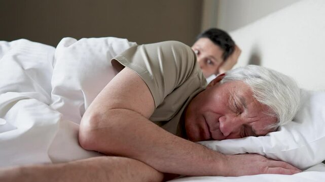 Closeup of elderly husband snoring disturbing wife sleep in bed. Angry stressed woman push and tries wake man, but no result. Sleep disruption, frustration, exhaustion, interrupted rest, helplessness