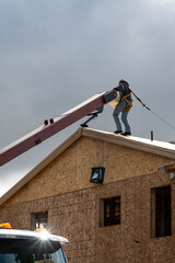 Workman in safety harness on wood framed house roof carrying package of roofing materials delivered by conveyor belt on a sunny winter day, home development construction project
