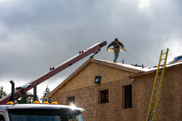 Workman in safety harness on wood framed house roof carrying package of roofing materials delivered by conveyor belt on a sunny winter day, home development construction project
