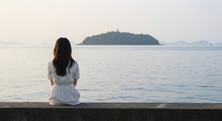 A young woman in a white dress sits on a concrete ledge by the sea, her back turned to the viewer