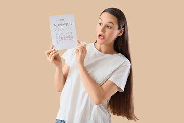 Surprised young woman with menstrual calendar on beige background