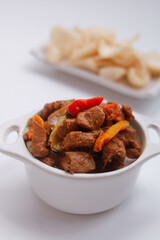 Close-up of a spicy chicken stir-fry in a white bowl with chili peppers and crackers in the background on a white surface