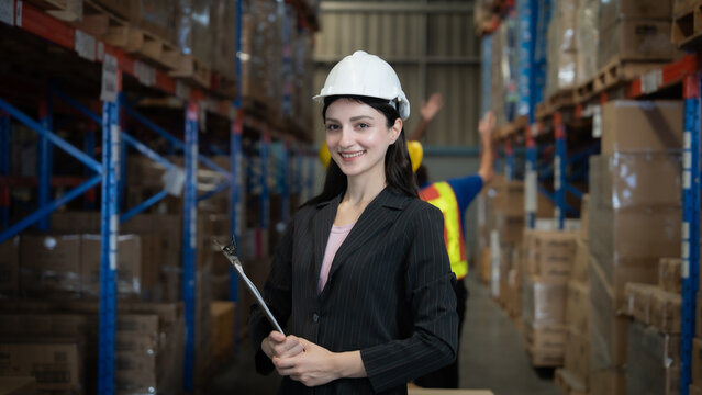 A woman wearing a hard hat and a suit is standing in a warehouse. She is holding a clipboard and smiling