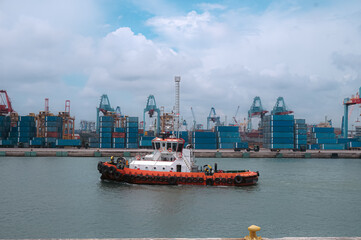 Fototapeta premium tugboat is centered in the water, moving slightly toward the right side of the image and navigating toward the port facilities.&nbsp;The mid-ground reveals a large container port.