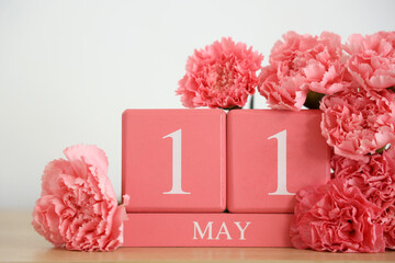 Cube calendar with date of Mother's Day and pink carnation flowers on shelf near light wall, closeup
