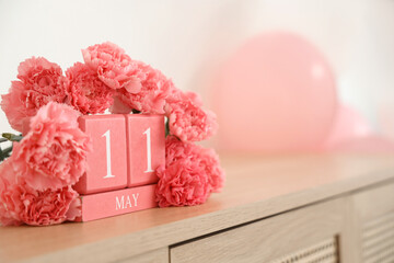 Cube calendar with date of Mother's Day and pink carnation flowers on shelf near light wall, closeup