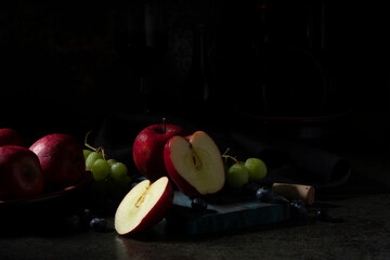 Red apples, green grapes, and blueberries on a chopping board with wine bottles in the dark background