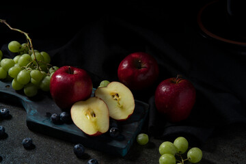 Close-up of a halved apple, red apples, green grapes, and blueberries on a chopping board on a black ceramic table