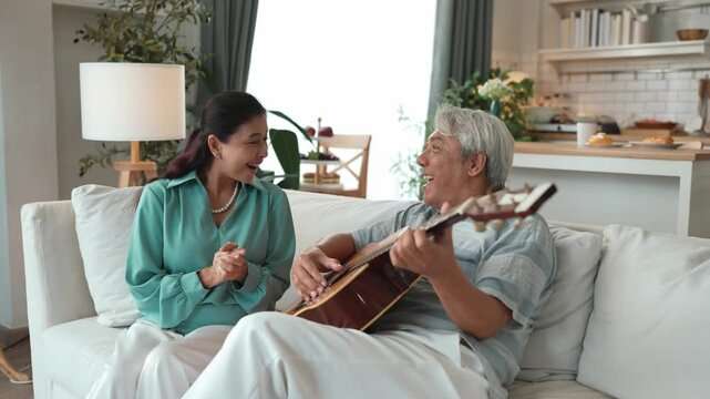 Happy senior man sitting at sofa and playing guitar or music instrument while his wife singing and moving to music. Elder couple spending time together and enjoy doing activity together. Myrmidon.