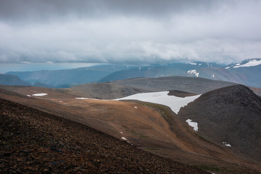 Dark overcast landscape with high pass, wide glacier on stony hill slope and snowy mountain range silhouette in far away in rainy low clouds. Large mountains with snow under gray sky in bad weather. - Powered by Adobe
