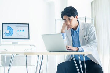 A young male doctor in a white coat uses a laptop in a medical office. A large screen in the background shows medical analytics and data visualizations, indicating a modern and technology-integrated h